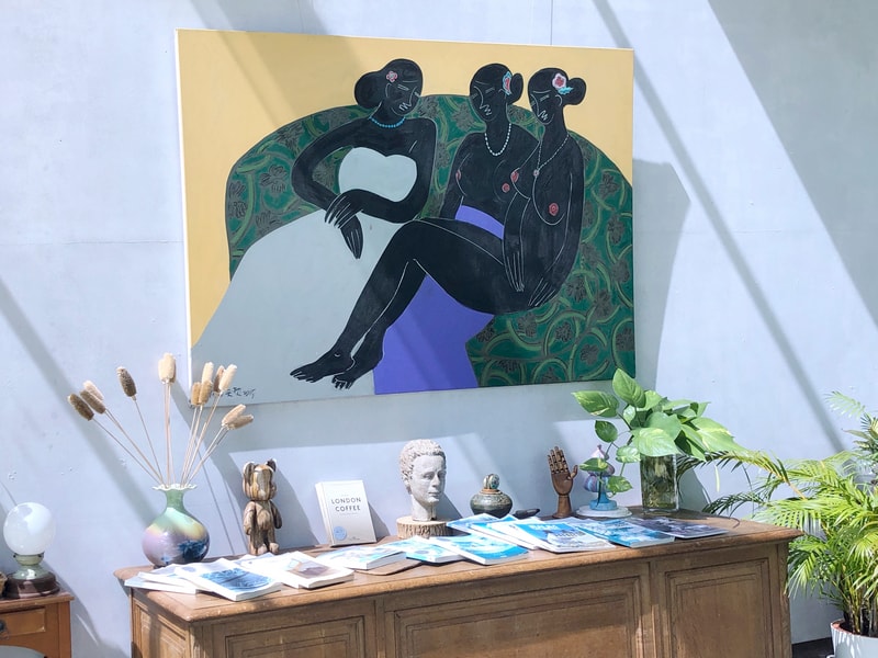 Arc Cafe 空間一隅，牆上畫作描繪三位人物，色彩濃烈，側櫃擺放乾燥植物、雕像與書籍，營造靜謐療癒氛圍。