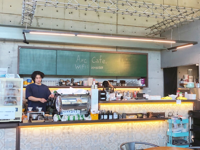 Arc Cafe 櫃檯區域，清水模牆面與綠色手寫黑板構成，空間寬敞明亮，設有兒童座椅與親子友善動線。