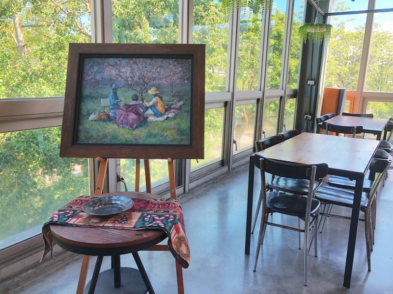 Arc Cafe 二樓空間，陽光透過玻璃灑入，木質桌椅沿窗排列，畫架上的畫作描繪三人野餐場景，營造靜謐與溫暖氛圍。