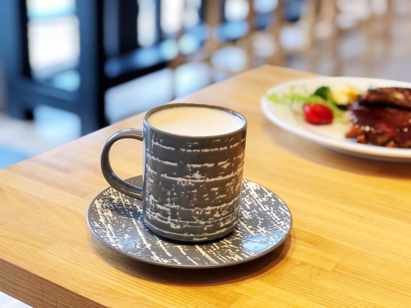 波赫士領地精品咖啡館明水店|台北大直不限時咖啡廳推薦,開心果奶霜千層必吃甜點 - 第29張圖 灰白紋理陶瓷杯盛裝英國伯爵鮮奶茶,杯盤相襯如霧中水墨,靜置於溫潤木桌上,背景點綴炙烤肉排與蔬菜,營造靜謐而細膩的午茶時光。